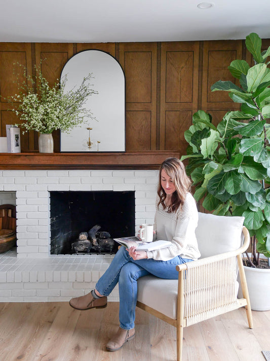 A woman sits in a chair by a fireplace, reading a book and holding a mug.