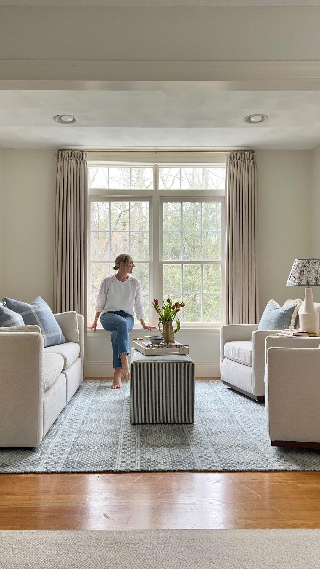 A woman sits on a windowsill in a bright living room with beige sofas