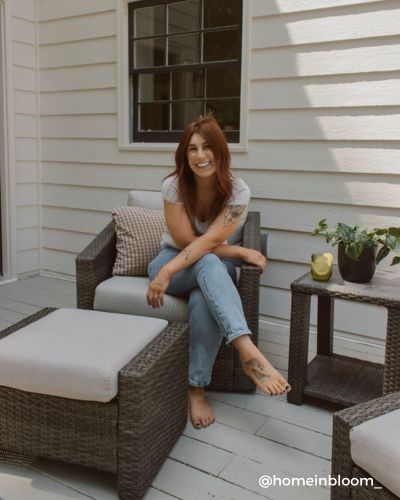 A woman with red hair and tattoos sits barefoot on a patio chair, smiling.