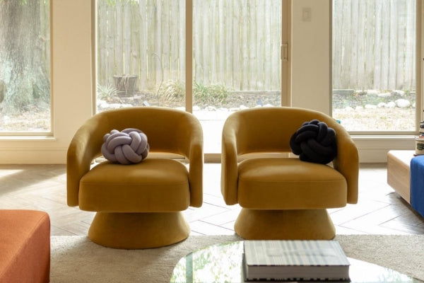 Two mustard-colored armchairs with knot pillows, in a bright room with large windows.