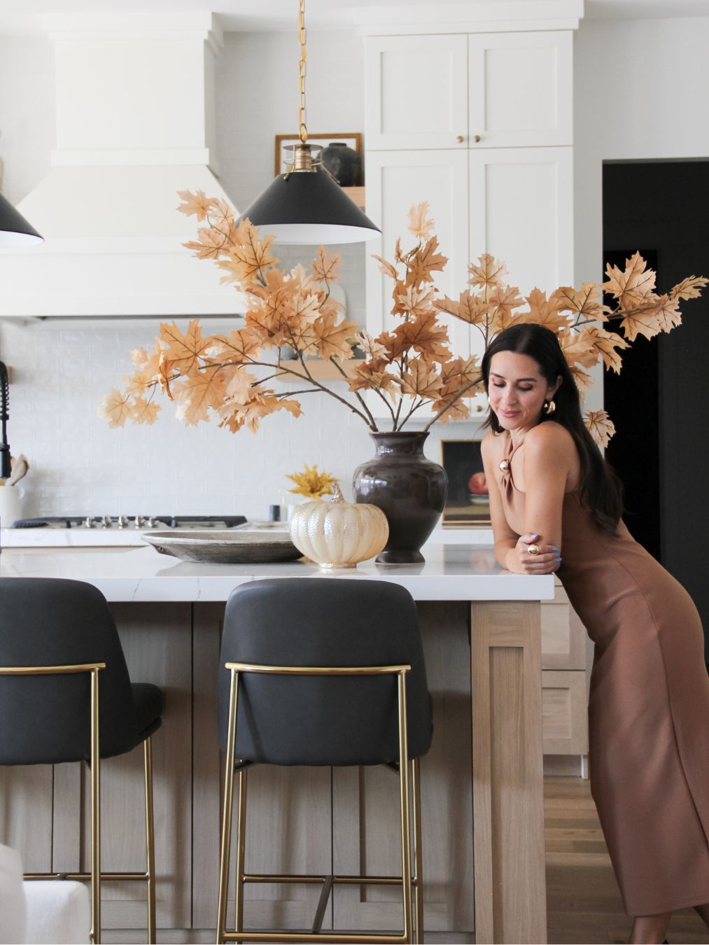 A woman in a brown dress leans on a kitchen island with black stools