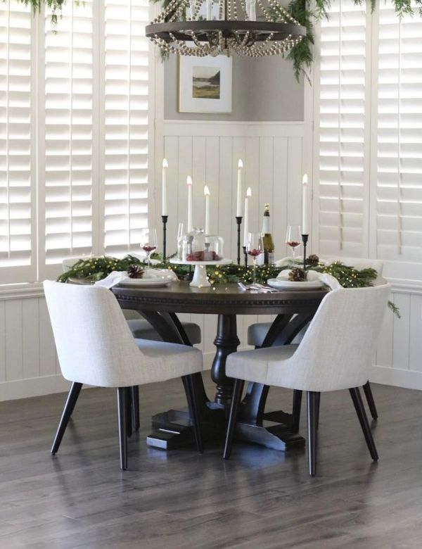 A festive dining table set with candles, greenery, and wine glasses, surrounded by white chairs.