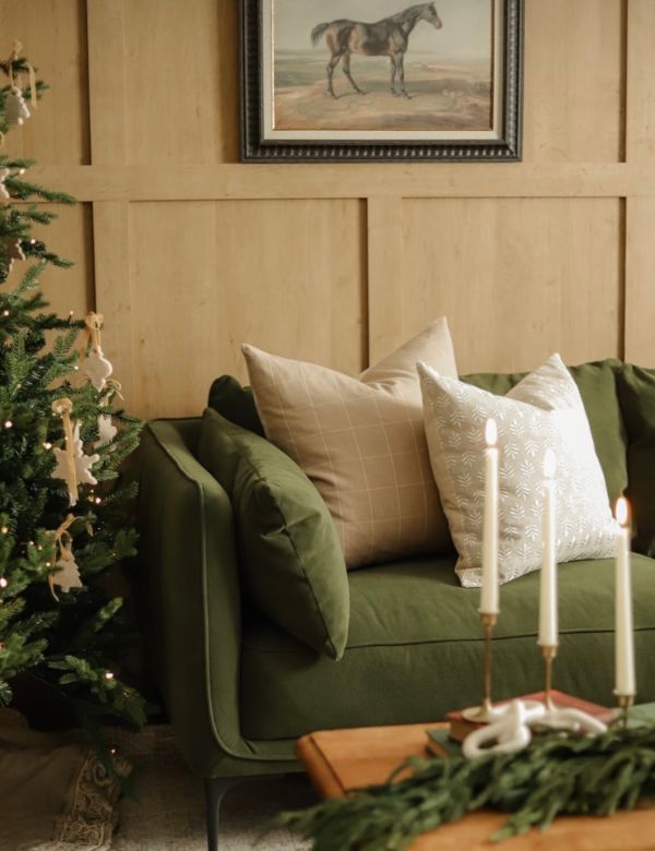 A cozy corner with a green sofa, white pillows, a lit candle, and a decorated Christmas tree.