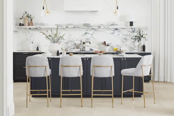 A modern kitchen with dark cabinets, a marble backsplash, and four gray upholstered bar stools with gold legs.