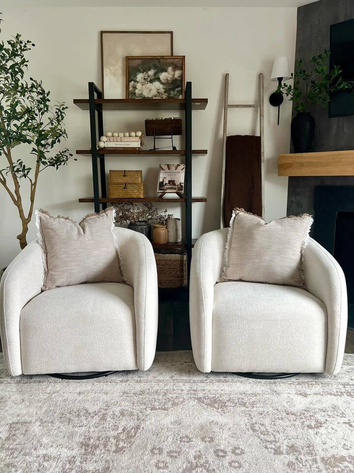 Two beige armchairs with textured pillows sit in front of a bookshelf filled with decorative items and a tall plant.